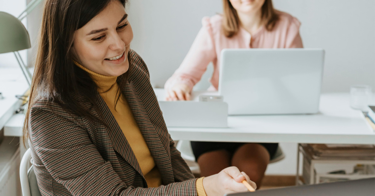 Kaksi naista istuu konttorissa. Edessä istuva nainen osoittaa takana istuvalle oman tietokoneensa näyttöä ja näyttää selittävän asiaa hymyillen.
